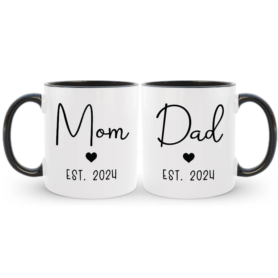 Two white mugs with black interior and handle, featuring 'Mom' and 'Dad' with heart symbols and 'Est. 2024' text.