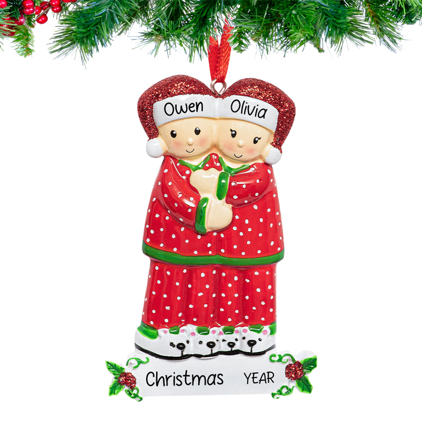 Personalized ornament of a couple in red Christmas pajamas with a heart above them, featuring custom names and year, hanging from a snow-dusted pine branch.