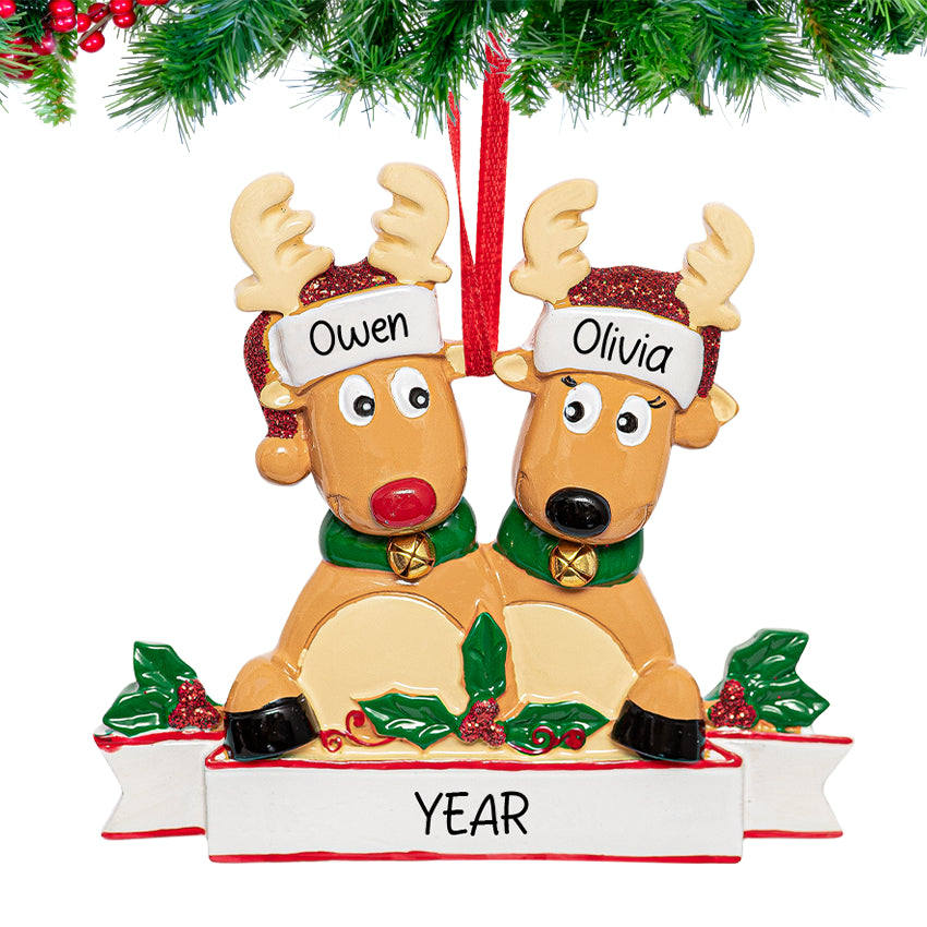 Two reindeer figurines nestled t, personalized with custom names and year, hanging from a snow-dusted pine branch.