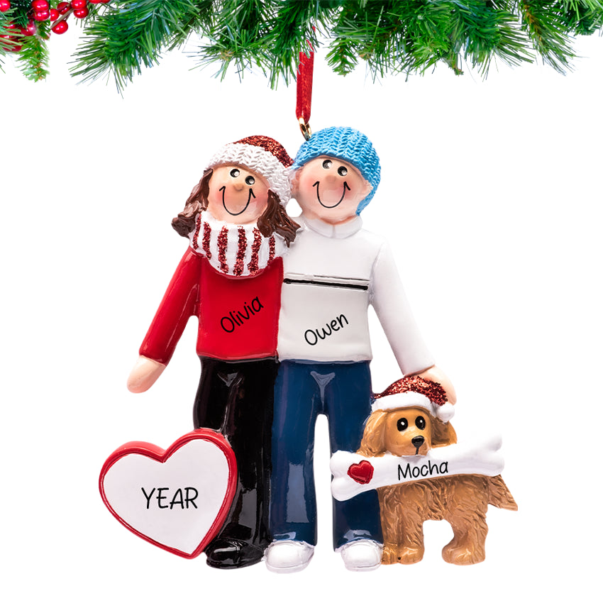 Couple holding their brown dog and smiling, names and 2025 engraved on a ornament hanging on a snow-dusted pine branch.