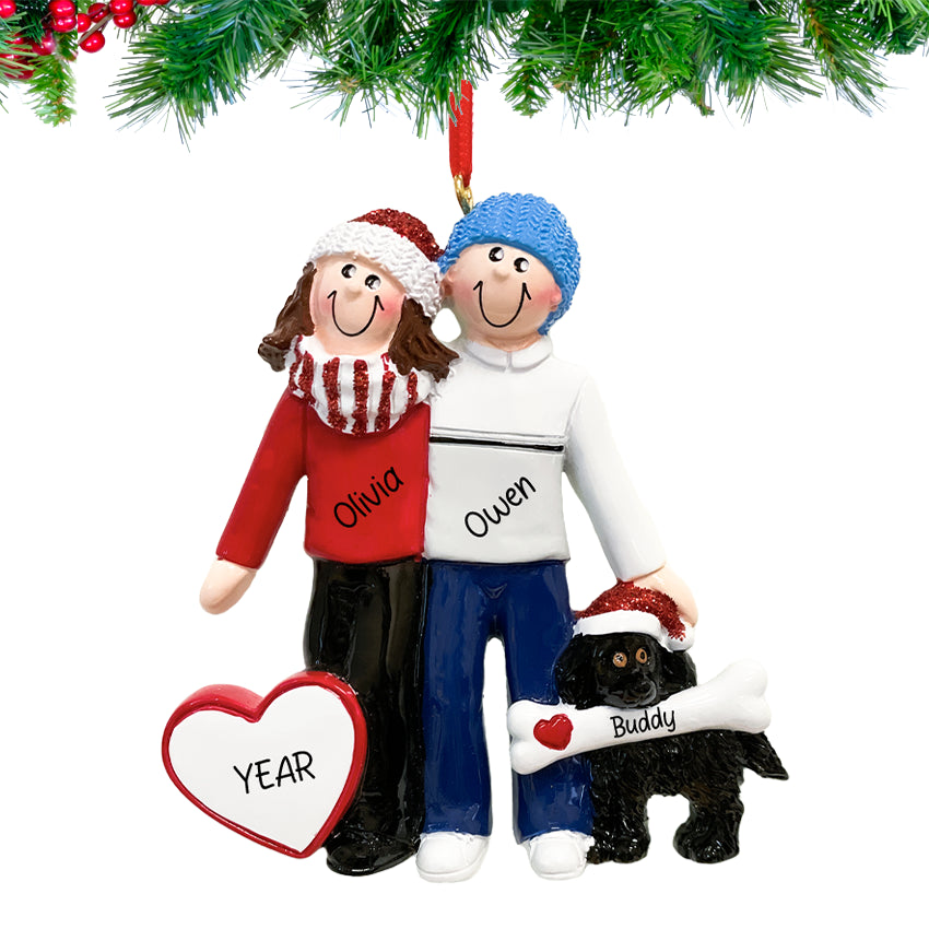Couple holding their brown dog and smiling, names and 2025 engraved on a ornament hanging on a snow-dusted pine branch.