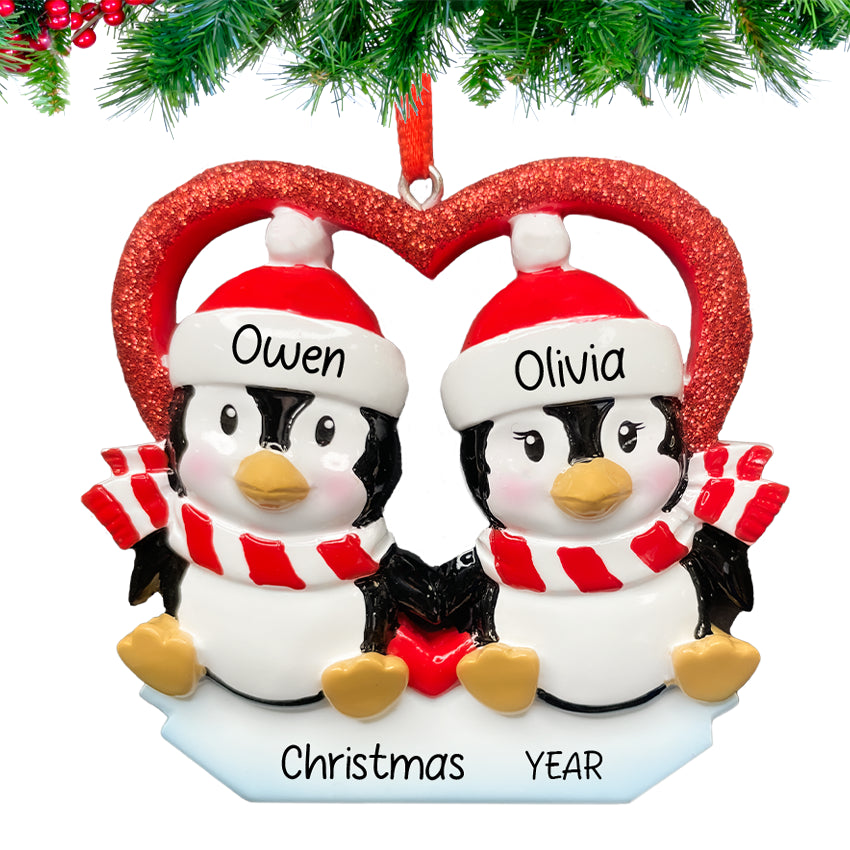 Two penguins sitting on a red heart, personalized with couple’s names and 2025, hanging on a snow-dusted Christmas tree branch.