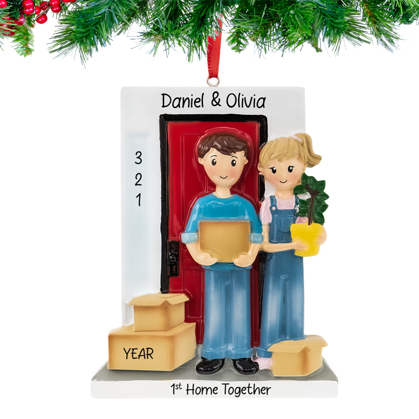 Happy couple holding a house key and moving boxes, personalized with names and 2025 on a Christmas ornament hanging from a festive tree branch.