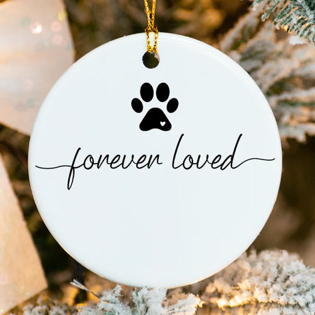Round white ornament with black paw print and 'forever loved' text, hanging on a tree.