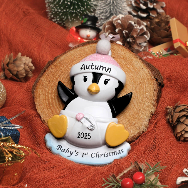 Penguin ornament with 'Autumn 2025 Baby's 1st Christmas' text on a red fabric background with pinecones and a small tree.