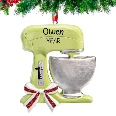“Stand mixer–shaped Christmas ornament hand-lettered with ‘[Name] [Year]’ hanging from a fir branch with a ribbon loop.”