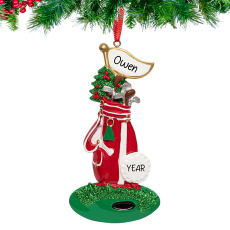 Stand bag and golf ball Christmas ornament engraved with ‘[Name] [Year]’ hanging from a snow-dusted fir branch with a ribbon loop.”